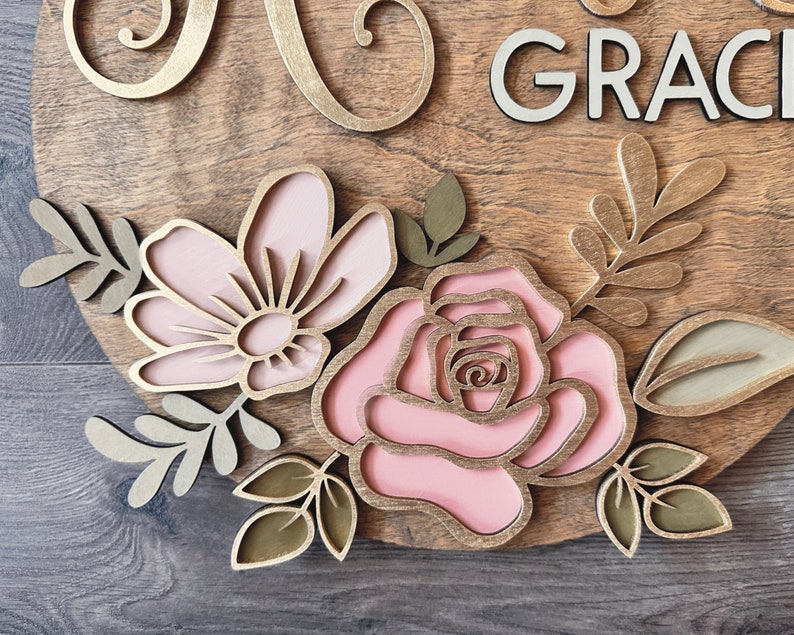 May include: A wooden sign with the name "Grace" in white letters and a floral design featuring pink and gold flowers and leaves.