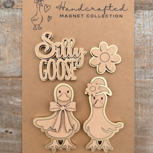 May include: A collection of handcrafted wooden magnets on a brown card. The magnets include the words "Silly Goose", a flower, and two goose figures. The card also features a goose illustration and the text "Handcrafted Magnet Collection".