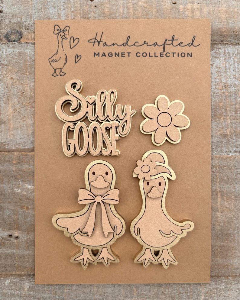 May include: A collection of handcrafted wooden magnets on a brown card. The magnets include the words "Silly Goose", a flower, and two goose figures. The card also features a goose illustration and the text "Handcrafted Magnet Collection".