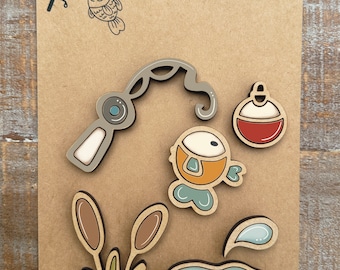 May include: A set of handcrafted fishing-themed magnets on a brown card. The magnets feature a fishing rod, fish, bobber, reeds, a pond, and a water droplet. The card is printed with the text "Handcrafted MAGNET COLLECTION".