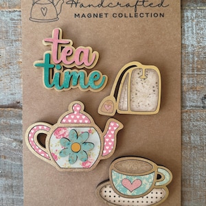 Puede incluir: Colección de imanes hechos a mano con una tetera, una taza de té, una bolsita de té y las palabras "Tea Time". La tetera tiene una tapa rosa con corazones y un diseño floral. La taza y el platillo tienen un diseño de corazón.