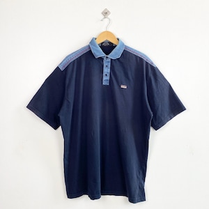 May include: A navy blue short-sleeve polo shirt featuring a light blue denim collar and shoulder panels with red stitching details. The shirt has a three-button placket and a small rectangular brand label on the left chest area.