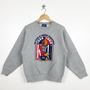 Pode incluir: Um moletom cinza com um design de basquete. O gráfico inclui uma bola de basquete, uma cesta e o texto "100 Years of Kansas Basketball 1898-1998", junto com um mascote Jayhawk e as letras "KU".