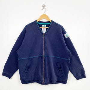 May include: Navy blue zip-up jacket with teal accents and a small logo patch. The jacket features a V-neck collar, two front pockets, and a full-length zipper. The Adidas logo is visible on the upper left sleeve.