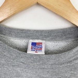 Op de afbeelding: Close-up van een grijze sweatshirt met een ronde hals. Het label toont de Amerikaanse vlag en de tekst "AMERICAN KNITWEAR MADE IN U.S.A. XL". De sweatshirt is gemaakt van een zacht, comfortabel materiaal.