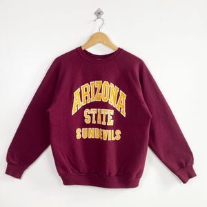 Puede incluir: Sudadera burdeos de cuello redondo con las palabras "ARIZONA STATE SUNDEVILS" en letras amarillas y blancas. La sudadera está colgada de una percha de madera sobre un fondo blanco.