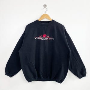 May include: Black crew neck sweatshirt with the text "WISCONSIN BADGERS" in red and white. The sweatshirt is hanging on a wooden hanger against a white background. The sweatshirt has long sleeves.