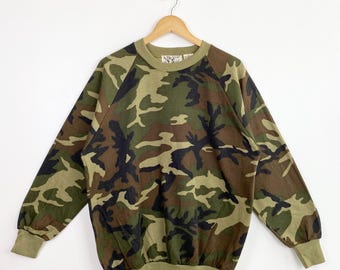 Könnte beinhalten: Ein Sweatshirt mit Camouflage-Muster in Grün-, Braun- und Schwarztönen. Das langärmlige Oberteil hat einen Rundhalsausschnitt und gerippte Bündchen und Saum. Das Sweatshirt hängt an einem Holzbügel vor weißem Hintergrund.