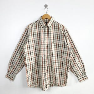 May include: A long-sleeve button-down shirt featuring a plaid pattern in shades of white, black, and rust orange. The shirt has a pointed collar, a front button placket, and a small embroidered logo on the left chest area, hanging on a wooden hanger.