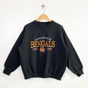 May include: Black crew neck sweatshirt featuring the text "Cincinnati Bengals" in orange and white. The design includes a football helmet graphic and the abbreviations "NFL" and "AFC". The sweatshirt is hanging on a wooden hanger.