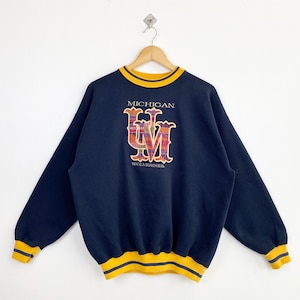 Peut inclure: Sweat-shirt bleu marine avec garniture jaune et marine. Le devant présente le mot "MICHIGAN" au-dessus d'un logo "UM" coloré et le mot "WOLVERINES" en dessous. Le sweat-shirt est suspendu à un cintre en bois.