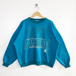 May include: Teal blue sweatshirt with the text "MIGHTY DUCKS OF ANAHEIM NATIONAL HOCKEY LEAGUE" in white and teal. The crew neck and long sleeves have teal cuffs and a teal hem. A vintage sports apparel item.