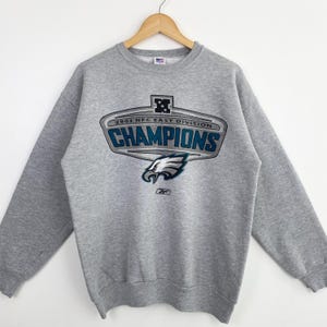 Op de afbeelding: Een grijze crewneck sweatshirt met een opdruk van "2002 NFC East Division CHAMPIONS". De afbeelding toont een adelaarskop en het Reebok-logo. De sweatshirt hangt aan een houten hanger.
