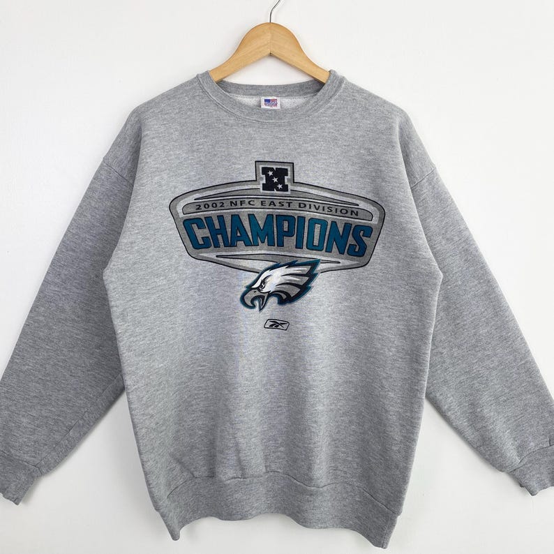 Op de afbeelding: Een grijze crewneck sweatshirt met een opdruk van "2002 NFC East Division CHAMPIONS". De afbeelding toont een adelaarskop en het Reebok-logo. De sweatshirt hangt aan een houten hanger.
