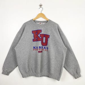 May include: A grey crew neck sweatshirt featuring the text "KU KANSAS FOOTBALL" in red and blue. The sweatshirt has long sleeves and a Nike logo. It is suspended from a wooden hanger.