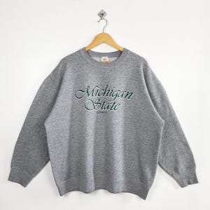 Puede incluir: Sudadera gris de cuello redondo con las palabras "Michigan State" bordadas en verde y blanco en el pecho. La sudadera está colgada en una percha de madera sobre un fondo blanco.