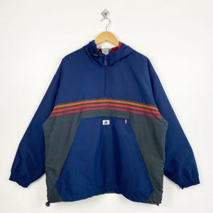May include: A navy blue Adidas anorak jacket with a hood and a half-zip closure. The jacket features three horizontal stripes in orange and red across the chest, and a small Adidas logo. The lower section is a darker blue and gray.