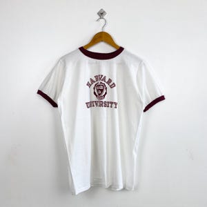 以下が含まれることがあります： 襟と袖にマルーンのトリムが付いた白いTシャツ。シャツの前面には、ハーバード大学のロゴがマルーンで表示されています。シャツは木製のハンガーに吊るされています。