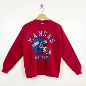 May include: A red sweatshirt featuring a blue football helmet with the word "KANSAS" arched above it in white lettering. Below the helmet, the word "JAYHAWKS" is displayed in a white box. The sweatshirt is hanging on a wooden hanger.