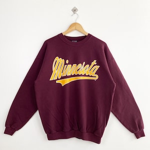 May include: A maroon sweatshirt with the word "Minnesota" in yellow script across the chest. The sweatshirt is hanging on a wooden hanger against a white background. The text has a yellow underline.