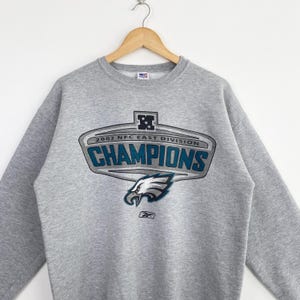 Op de afbeelding: Een grijze sweatshirt met een afbeelding met de tekst "2002 NFC EAST DIVISION CHAMPIONS" in teal en wit. De afbeelding bevat een adelaarskop en het Reebok-logo. De sweatshirt hangt aan een houten hanger.