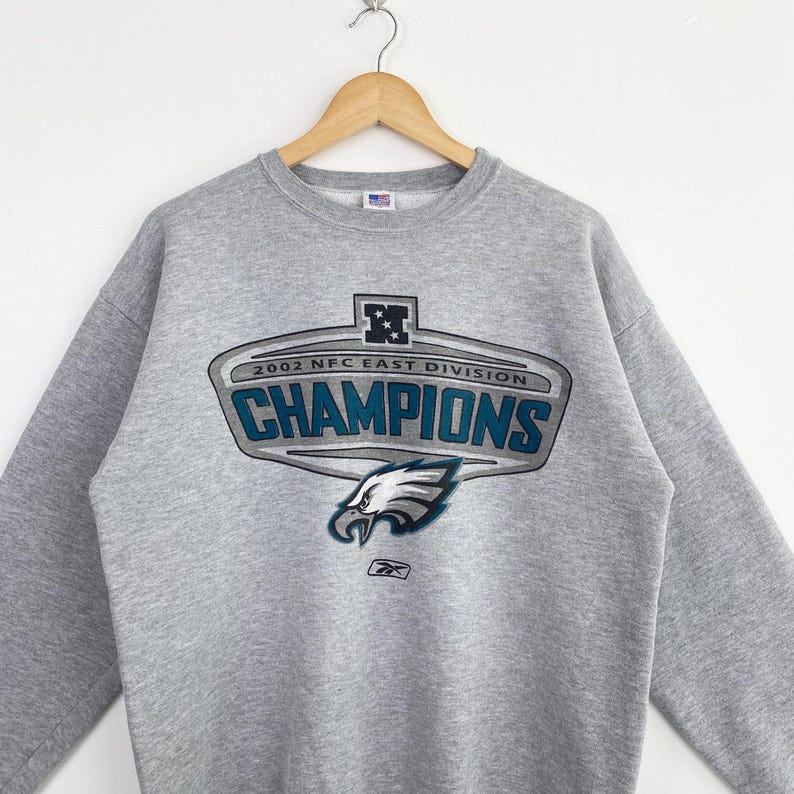 Op de afbeelding: Een grijze sweatshirt met een afbeelding met de tekst "2002 NFC EAST DIVISION CHAMPIONS" in teal en wit. De afbeelding bevat een adelaarskop en het Reebok-logo. De sweatshirt hangt aan een houten hanger.