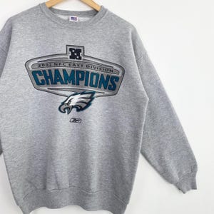 Op de afbeelding: Een grijze sweatshirt met een ronde hals en lange mouwen. De voorkant heeft een afbeelding met de tekst "2002 NFC EAST DIVISION CHAMPIONS" en een adelaarslogo. Het Reebok-logo staat onderaan.