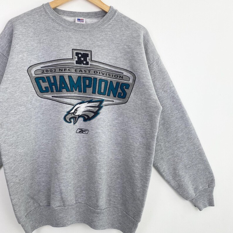 Op de afbeelding: Een grijze sweatshirt met een ronde hals en lange mouwen. De voorkant heeft een afbeelding met de tekst "2002 NFC EAST DIVISION CHAMPIONS" en een adelaarslogo. Het Reebok-logo staat onderaan.