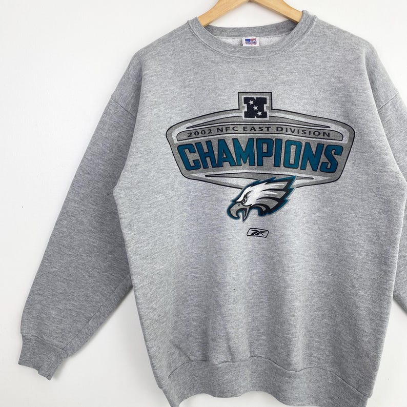 Op de afbeelding: Een grijze sweatshirt met de tekst "2002 NFC East Division Champions" en een adelaarslogo. De sweatshirt met lange mouwen is gemaakt van zachte stof. Het Reebok-logo is ook zichtbaar.