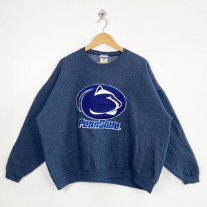 May include: Navy blue sweatshirt with the Penn State logo in white and blue. The logo features a stylized Nittany Lion head inside a blue oval with the words "Penn State" below it. The sweatshirt is hanging on a wooden hanger.