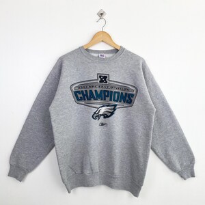 Op de afbeelding: Een grijze sweatshirt met de tekst "CHAMPIONS" in blauw en wit, samen met de woorden "2002 NFC EAST DIVISION". De sweatshirt heeft een teamlogo en het Reebok-logo.