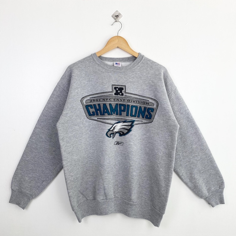 Op de afbeelding: Een grijze sweatshirt met de tekst "CHAMPIONS" in blauw en wit, samen met de woorden "2002 NFC EAST DIVISION". De sweatshirt heeft een teamlogo en het Reebok-logo.