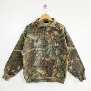 Könnte beinhalten: Ein Sweatshirt mit Camouflage-Muster, Viertelreißverschluss und Kängurutasche. Das Design integriert Braun-, Grün- und Beigetöne, die eine Waldumgebung nachahmen. Das langärmlige Oberteil hängt an einem Holzbügel.