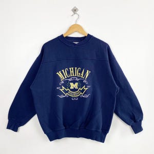 May include: Navy blue sweatshirt with the word "MICHIGAN" arched across the chest in yellow script. Below the word "MICHIGAN" is a yellow "M" logo with the word "WOLVERINES" below it. The sweatshirt has a crew neck and long sleeves.