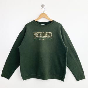 May include: A dark green sweatshirt with the text "NORTH DAKOTA STATE UNIVERSITY ALUMNI" in gold lettering. The sweatshirt is hanging on a wooden hanger against a white background. The garment has long sleeves and a crew neck.