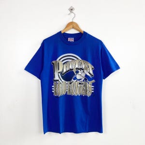 May include: A royal blue short-sleeve t-shirt featuring a graphic print of a cartoon bird holding a hockey stick. The text PHOENIX ROADRUNNERS is printed in a metallic gold font over a circular black and white target design on the chest.