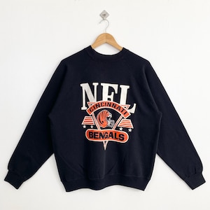May include: A navy blue sweatshirt featuring the NFL logo with "Cincinnati Bengals" in orange and white. The design includes a football helmet and stars. The sweatshirt is hanging on a wooden hanger against a white background.
