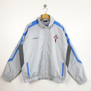 May include: A light gray zip-up jacket with blue and gray accents. The jacket features a small logo on the chest and a team emblem. The jacket has a stand-up collar and long sleeves. The jacket is hanging on a wooden hanger.
