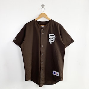 Può includere: Una maglia da baseball marrone con il logo bianco dei San Francisco Giants sul davanti. La maglia ha maniche corte, una chiusura con bottoni e una piccola toppa in basso a destra. È appesa a una gruccia di legno.