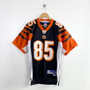 May include: Black and orange football jersey with the number 85 in white. The jersey has orange and black stripes on the sleeves and the team logo on the chest. The jersey is hanging on a wooden hanger.