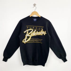 May include: Black Purdue Boilermakers sweatshirt with gold and white lettering. The sweatshirt features the words "PURDUE", "Boilermakers", and "UNIVERSITY" in a stylized font. The long-sleeved crew neck sweatshirt is hanging on a wooden hanger.