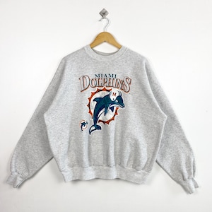 Puede incluir: Sudadera gris claro con el logotipo de los Miami Dolphins. El logotipo muestra un delfín azul saltando a través de un diseño de sol, con el nombre del equipo arqueado arriba. La sudadera tiene mangas largas y cuello redondo.