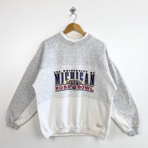 May include: A heather grey and white colour-blocked crewneck sweatshirt featuring the text THE UNIVERSITY OF MICHIGAN 1993 ROSE BOWL with a rose graphic in the centre. The long-sleeved garment has a ribbed collar, cuffs, and hem.