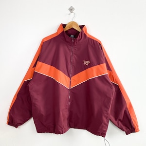 May include: A maroon and orange zip-up jacket with a white accent stripe. The jacket features a chevron design across the chest and sleeves. The letters "VT" are embroidered on the chest. The jacket is hanging on a wooden hanger.