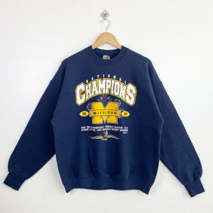 以下が含まれることがあります： 白と金色の文字で「NATIONAL CHAMPIONS」と書かれたネイビーブルーのスウェットシャツ。 金色の「MICHIGAN」ロゴとテキスト「BIG TEN CHAMPIONS PERFECT SEASON 12-0 HOME OF THE 1997 HEISMAN TROPHY WINNER」が特徴です。