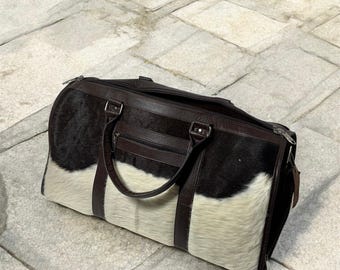 May include: A large, rectangular duffel bag with a combination of black and white cowhide and brown leather accents. The bag features a top zipper closure, a front zipper pocket, and two handles. The bag is designed for travel.