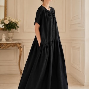 Peut inclure: Une robe longue noire à manches courtes, dotée d'une encolure ronde et d'une patte de boutonnage sur le devant. Le vêtement présente une taille froncée créant une jupe ample et fluide avec des poches latérales, portée avec des sandales noires.