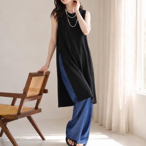 May include: A sleeveless black tunic with side slits, paired with wide-leg blue jeans. The outfit is accessorized with a layered necklace. A wooden chair is visible on the left side of the image.