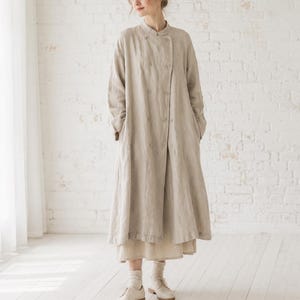 May include: A long, beige, double-breasted coat with a mandarin collar and long sleeves. The coat features two vertical rows of matching buttons down the front, a loose, flowing silhouette, and is worn over a matching beige skirt and light-colored shoes.