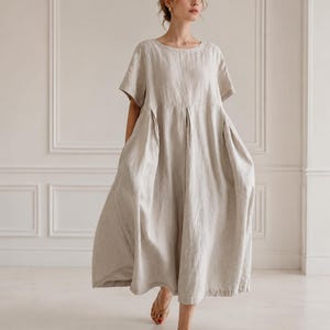 May include: A beige, loose-fitting, short-sleeved midi dress made of a textured fabric. The dress features a round neckline, a high waistline with subtle pleats that create a voluminous skirt, and a hemline that falls below the knees.